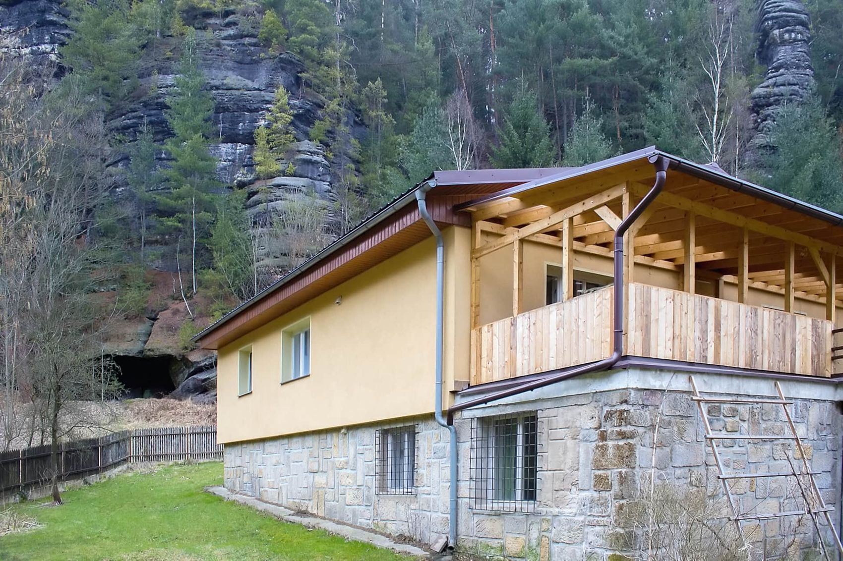 Dům se žlutou fasádou a dřevěnou terasou pod pískovcovou skalní stěnou, vlevo vstup do jeskyně
